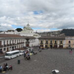 An elevated view of the historic Plaza San Francisco in Quito, Ecuador, featuring a wide stone courtyard surrounded by yellow and white colonial buildings under a cloudy sky.