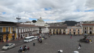 An elevated view of the historic Plaza San Francisco in Quito, Ecuador, featuring a wide stone courtyard surrounded by yellow and white colonial buildings under a cloudy sky.