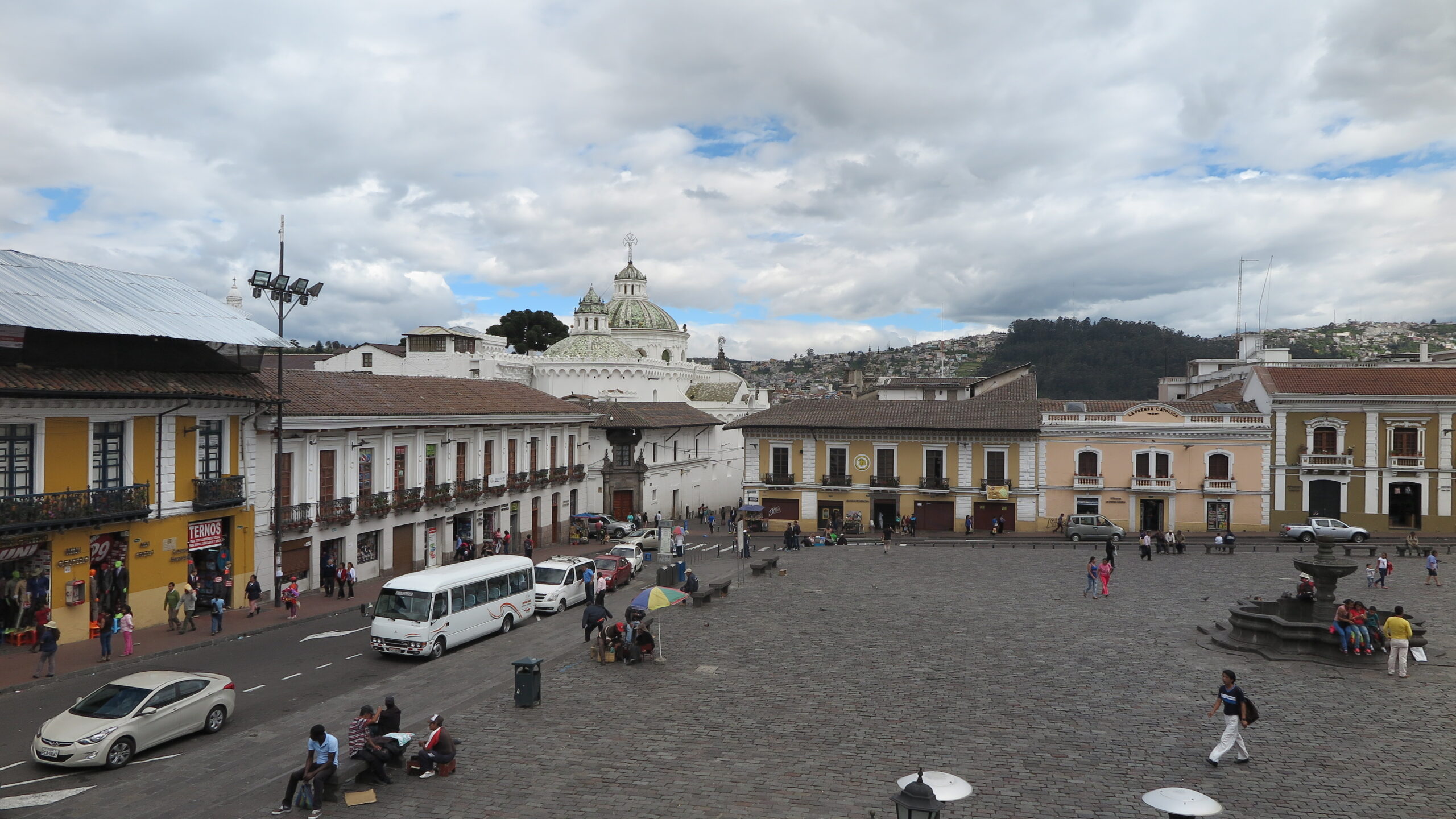An elevated view of the historic Plaza San Francisco in Quito, Ecuador, featuring a wide stone courtyard surrounded by yellow and white colonial buildings under a cloudy sky.