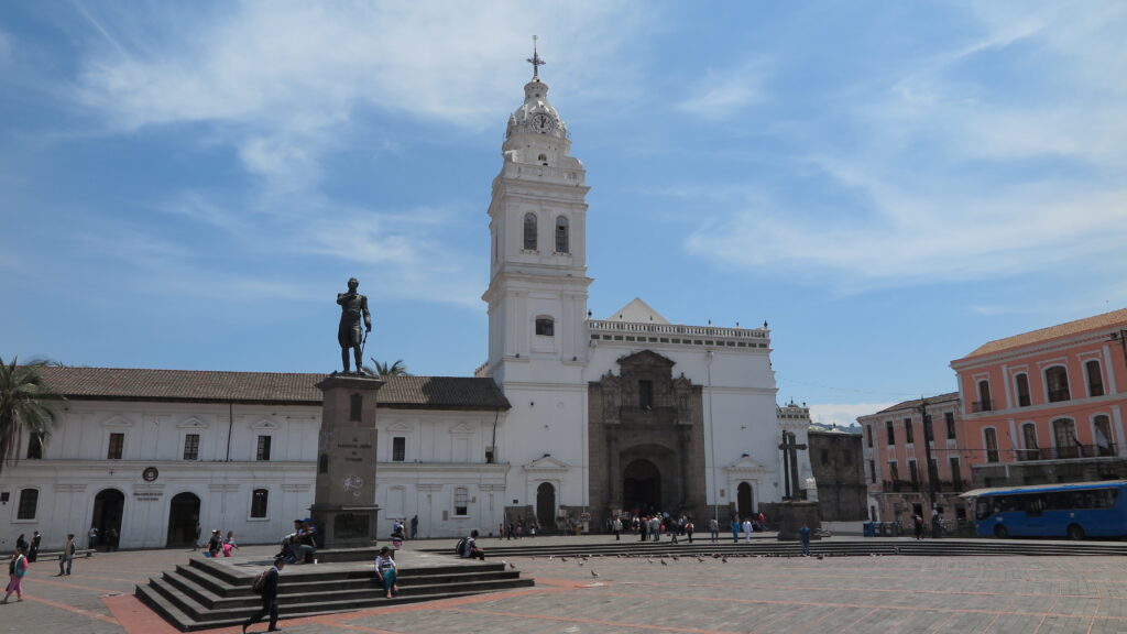 The wide-open Plaza de Santo Domingo in the historic center of Quito, Ecuador, featuring the prominent statue of Antonio José de Sucre and the majestic white Iglesia de Santo Domingo under a blue sky.