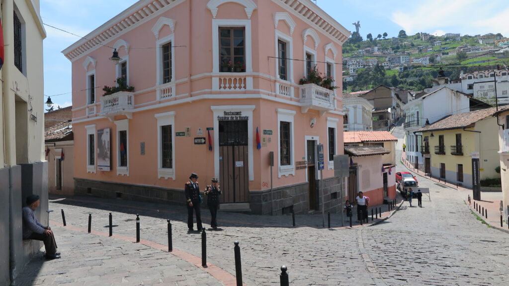 A cobblestone street corner in historic Quito, Ecuador, featuring a prominent two-story salmon pink colonial building and officers walking, with El Panecillo hill visible in the background.