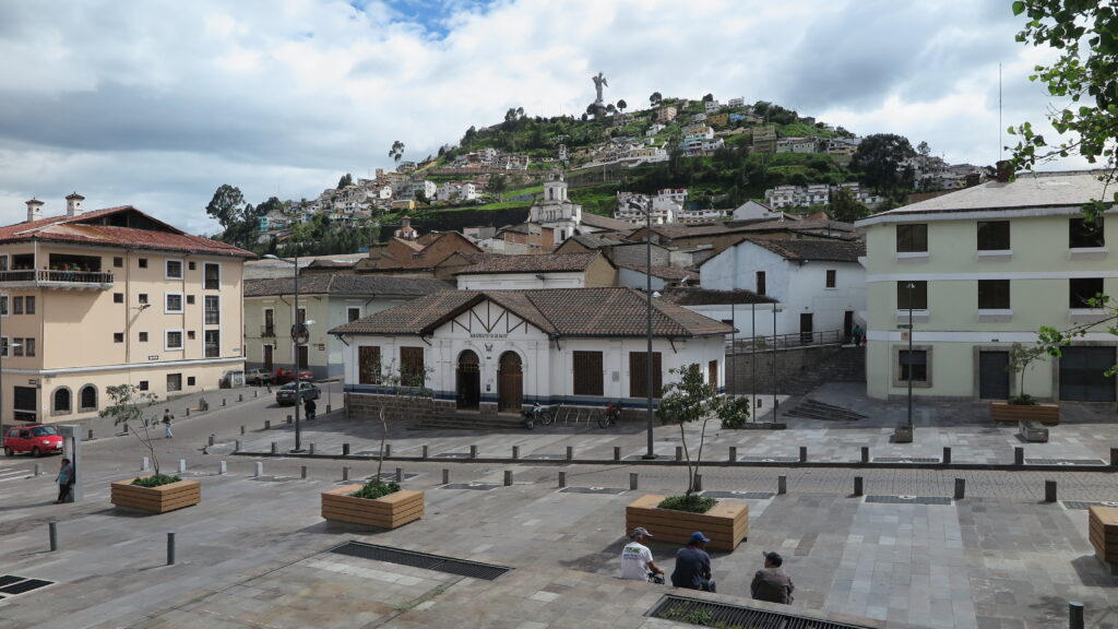 A scenic view of El Panecillo hill in Quito, Ecuador, topped with the giant winged Virgen del Panecillo statue, overlooking a historic plaza and colonial houses.