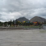 A wide concrete plaza at the Mitad del Mundo (Equator Line) complex in Quito, Ecuador, lined with international flags and framed by Andean mountain peaks under an overcast sky.