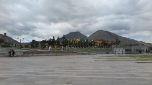 A wide concrete plaza at the Mitad del Mundo (Equator Line) complex in Quito, Ecuador, lined with international flags and framed by Andean mountain peaks under an overcast sky.