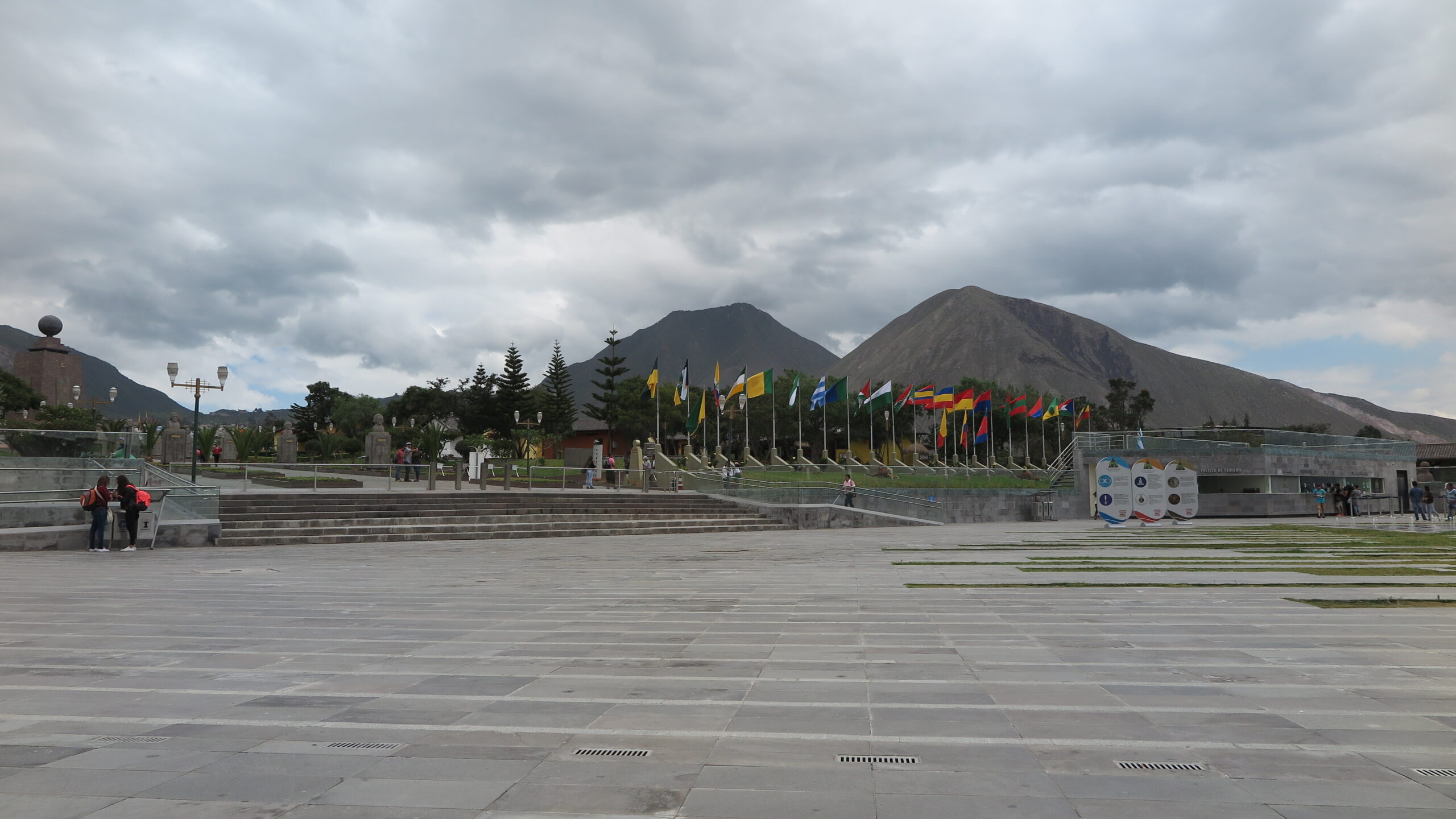 A wide concrete plaza at the Mitad del Mundo (Equator Line) complex in Quito, Ecuador, lined with international flags and framed by Andean mountain peaks under an overcast sky.
