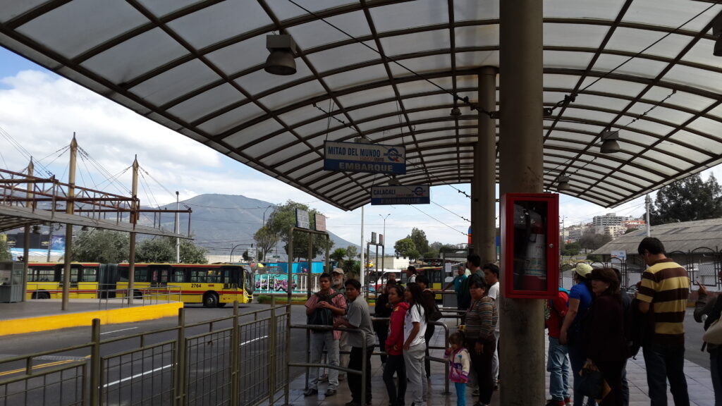 Passengers waiting under a covered transit platform at Terminal Ofelia in Quito, Ecuador, with a blue sign pointing to the Mitad del Mundo (Equator Line) bus boarding area.