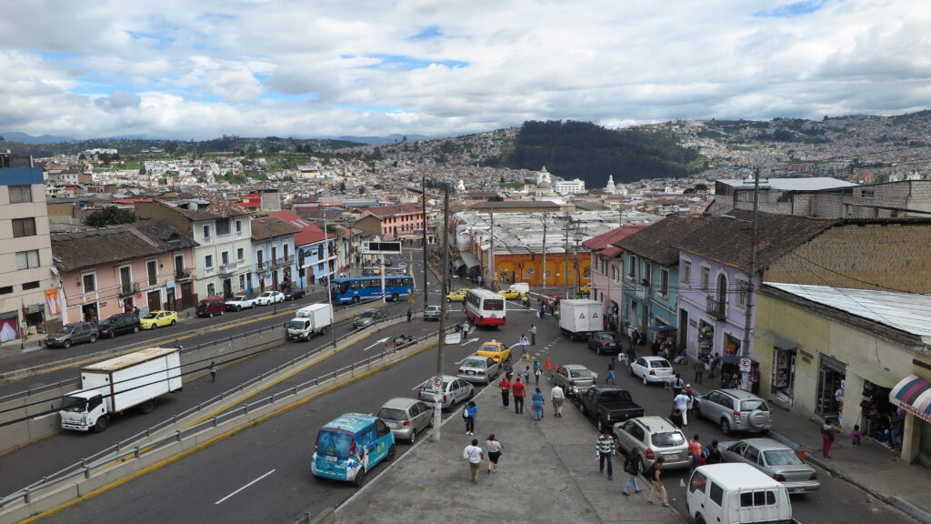 An elevated, wide-angle shot of a busy street intersection in Quito, Ecuador, featuring colorful colonial buildings, local traffic, pedestrians, and sprawling hills with houses in the background under a cloudy sky.