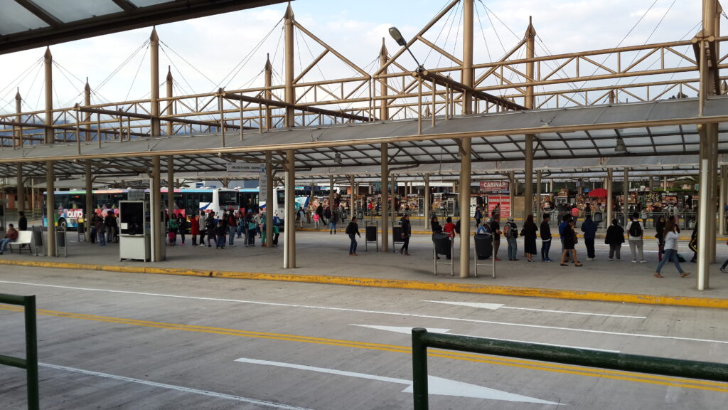 People waiting at the outdoor platforms of a busy bus terminal in Quito, Ecuador, under a large metal canopy structure.