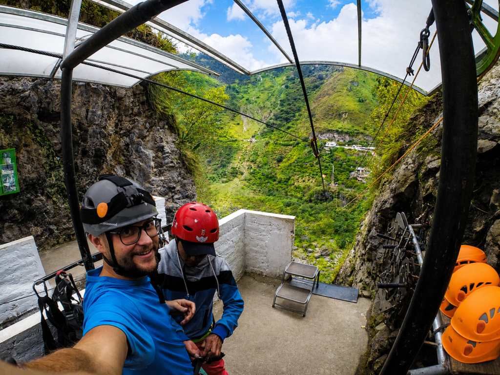 Ecuador Baños zipline istasyonunda, mavi tişörtlü ve kasklı bir turistin gülümseyerek çektiği selfie. Görevli emniyet kemerini kontrol ederken, arka planda yemyeşil dik dağlar ve derin bir vadi görünüyor.