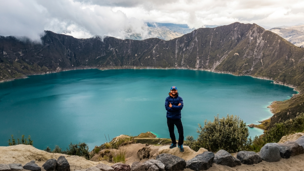 Ecuador Laguna de Quilotoa A full-body photograph of a smiling man wearing a blue jacket and hat, standing with arms crossed at a viewpoint overlooking the massive, vibrant turquoise crater lake of the Quilotoa volcano in Ecuador. Rugged crater walls and a cloudy sky form the background. Ecuador Laguna de Quilotoa!