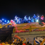 A night photograph taken in Cali, Colombia, shows a carnival float in a parade on a street. On the float, which is designed with large glowing instrument sculptures like a trumpet and a saxophone, a live salsa band performs under spotlights. A dense crowd on the street below watches the float, some holding up smartphones to take photos, and one person raises a flag. The float is decorated with the text "Feria de Cali."
