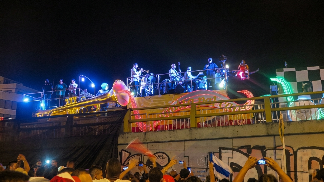 A night photograph taken in Cali, Colombia, shows a carnival float in a parade on a street. On the float, which is designed with large glowing instrument sculptures like a trumpet and a saxophone, a live salsa band performs under spotlights. A dense crowd on the street below watches the float, some holding up smartphones to take photos, and one person raises a flag. The float is decorated with the text "Feria de Cali."