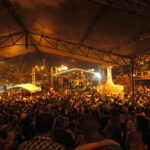 A wide night shot of a massive crowd gathered under a large temporary tent structure during the Feria de Cali in Cali, Colombia. In the background, an illuminated outdoor concert stage and a illuminated statue are visible as people enjoy live music and dance salsa in Cali.
