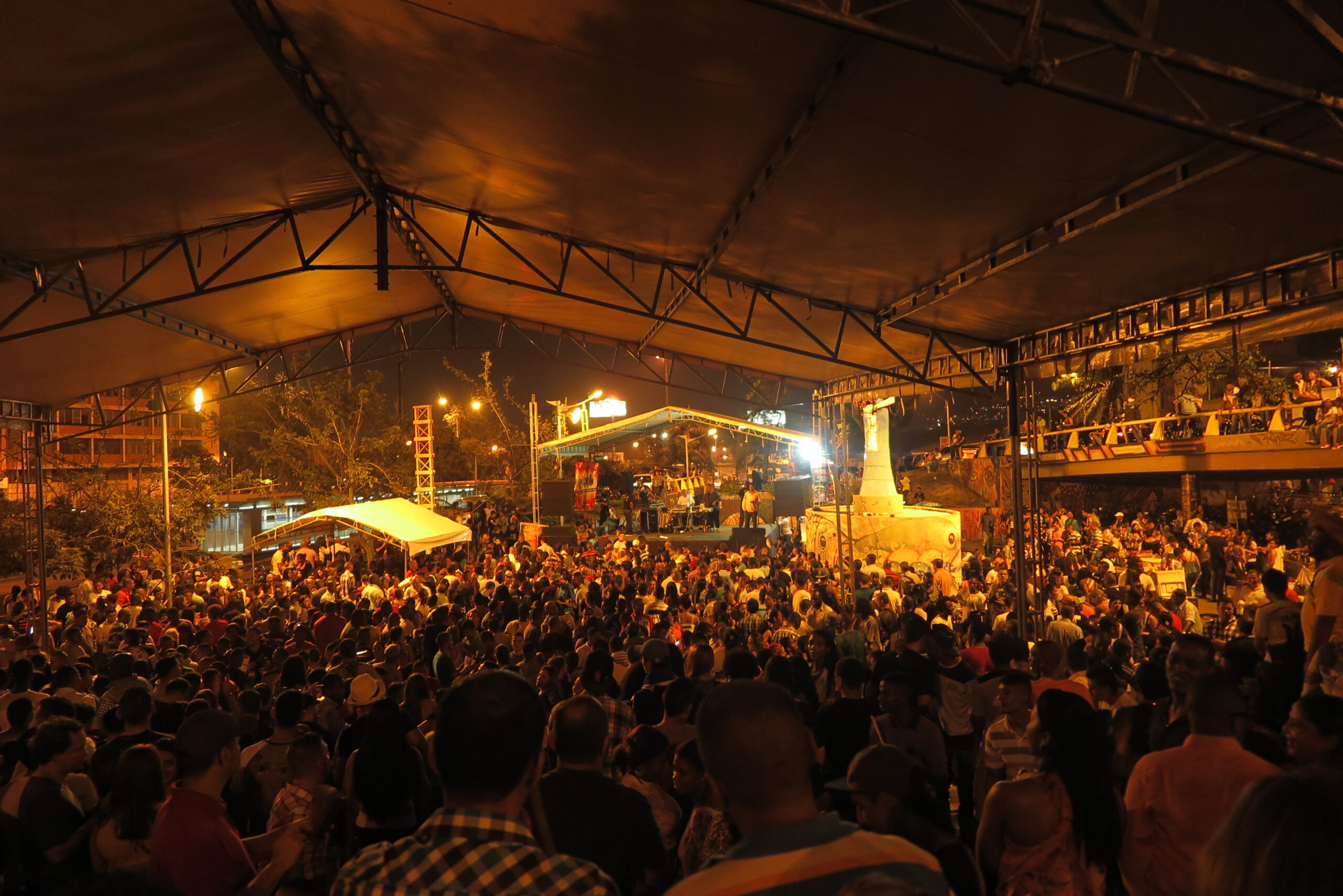 A wide night shot of a massive crowd gathered under a large temporary tent structure during the Feria de Cali in Cali, Colombia. In the background, an illuminated outdoor concert stage and a illuminated statue are visible as people enjoy live music and dance salsa in Cali.