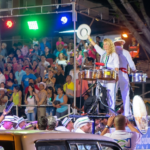 A night photograph of a live performance on an elevated float during the Feria de Cali in Cali, Colombia. In the foreground, a woman in a white uniform plays timbales and waves a white hat, with colored stage lights behind her and a large crowd looking on, celebrating salsa in Cali.