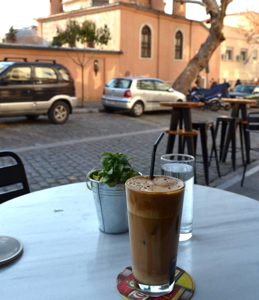 1901 Kavala Greece Where to eat in Kavala - A close-up view from a cafe table featuring a tall glass of iced coffee (frappe) and a glass of water on a cobblestone street in Kavala, Greece, with modern cars and the historical landmark, the Imaret, visible in the background.