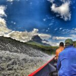A point-of-view photograph taken from inside a wooden boat, with water splashing in the foreground, showing two men looking ahead at a lush, forested mountain under a dramatically cloudy blue sky.