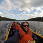 Angel Falls - A cheerful group selfie taken in a wooden boat on a river, featuring a smiling man with a beard and sunglasses in an orange life vest in the foreground, with other passengers, lush forested riverbanks, and a cloudy sky.