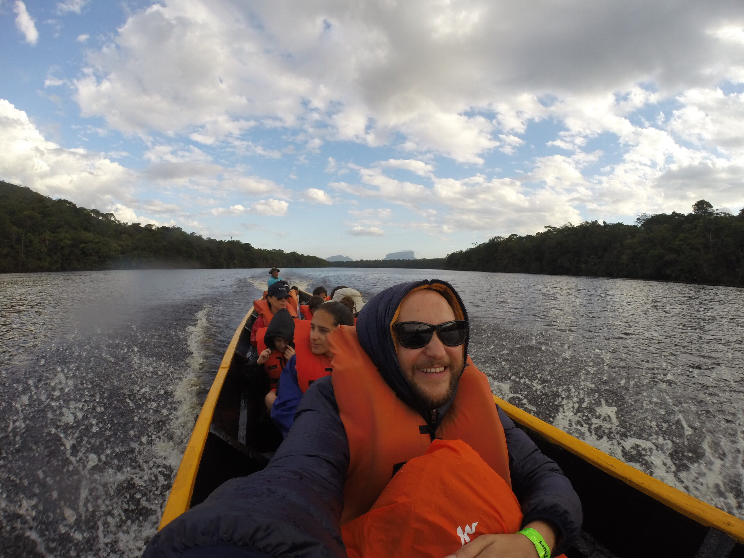 Angel Falls - A cheerful group selfie taken in a wooden boat on a river, featuring a smiling man with a beard and sunglasses in an orange life vest in the foreground, with other passengers, lush forested riverbanks, and a cloudy sky.