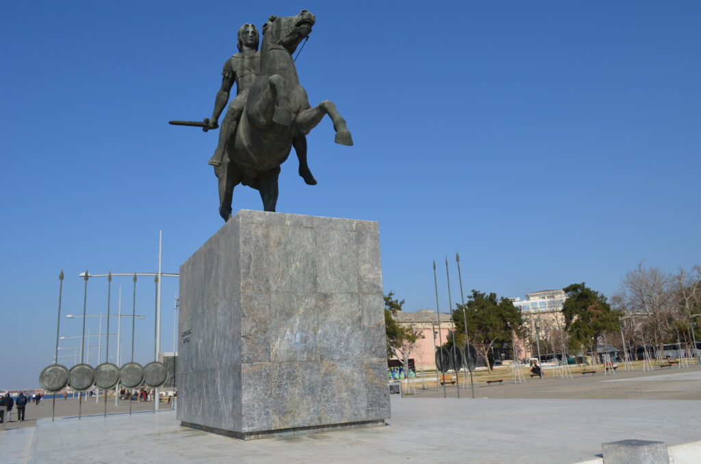 Bronze equestrian statue of Alexander the Great on a tall marble pedestal at the Thessaloniki waterfront, with shields and spears beside it.