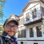 A traveler wearing glasses and a cap taking a smiling selfie in front of the Atatürk House Museum in Thessaloniki.