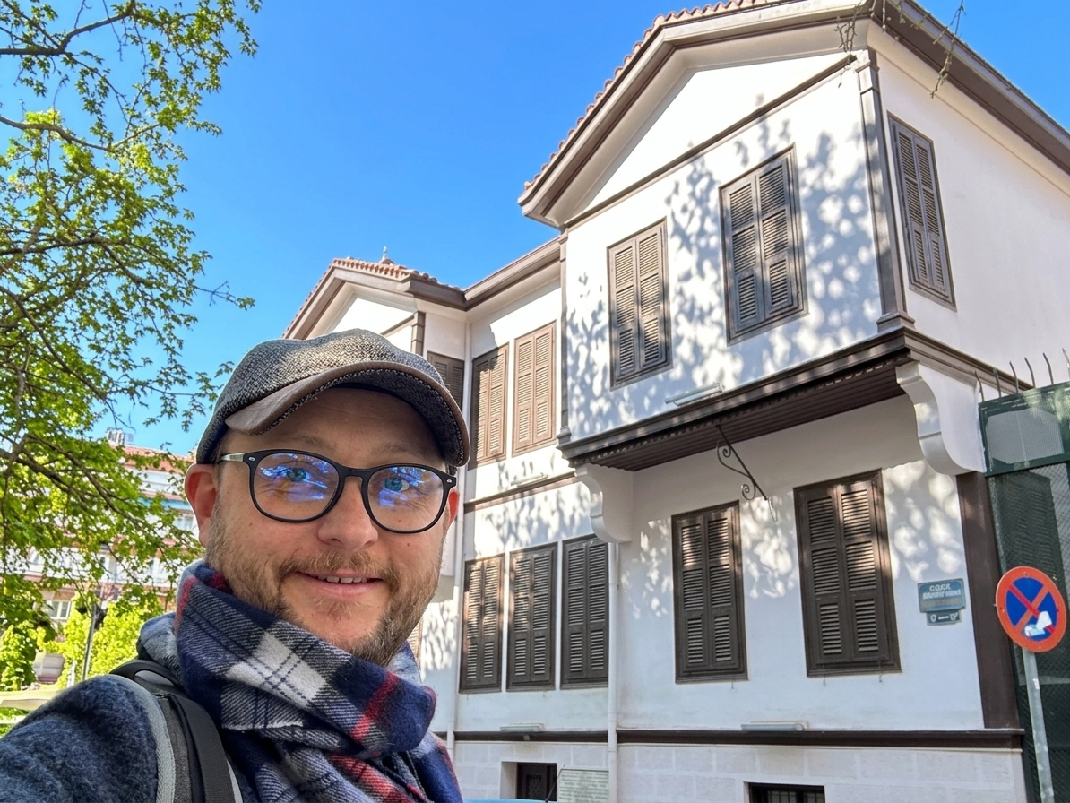 A traveler wearing glasses and a cap taking a smiling selfie in front of the Atatürk House Museum in Thessaloniki.