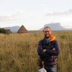 Angel Falls - A smiling man standing in a tall grassy field with indigenous thatched-roof huts and iconic flat-topped tepui mountains in the background at Canaima National Park, Venezuela.
