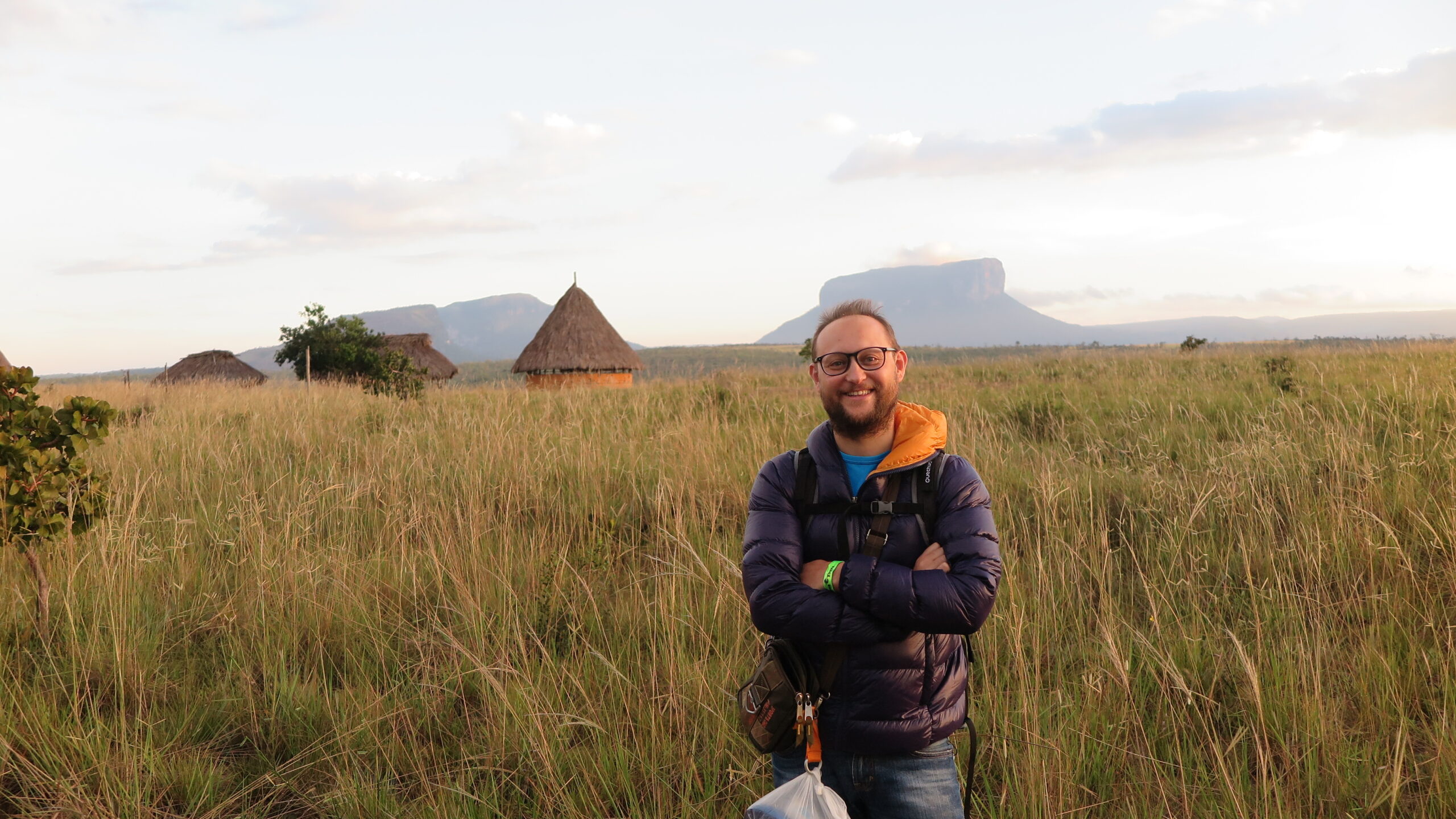 Angel Falls - A smiling man standing in a tall grassy field with indigenous thatched-roof huts and iconic flat-topped tepui mountains in the background at Canaima National Park, Venezuela.