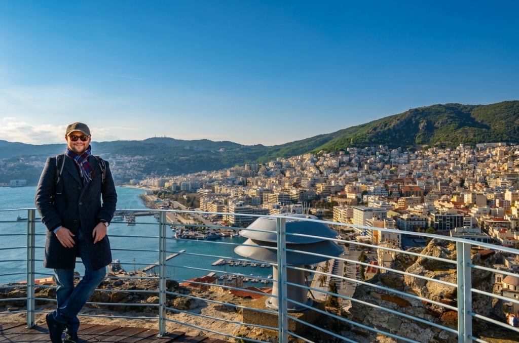 Things to do in Kavala - A panoramic photograph taken from the fortified stone ramparts of Kavala Castle in Greece. On the left, a smiling man in a dark coat, flat cap, scarf, and sunglasses leans on a white metal railing, with one leg crossed over the other. The background is a vast cityscape of Kavala, with white buildings descending a steep hillside to a bustling harbor and marina filled with boats. Beyond the bay are lush, tree-covered mountains under a clear blue sky. A circular metal vent is in the lower right foreground.