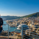 Kavala'da Gezilecek Yerler - Yunanistan'daki Kavala Kalesi'nin taş surlarından çekilmiş panoramik bir fotoğraf. Solda, koyu renkli palto, kasket, atkı ve güneş gözlüğü takan gülümseyen bir adam, bir bacağını diğerinin üzerine atmış şekilde beyaz metal bir korkuluğa yaslanıyor. Arka planda, teknelerle dolu hareketli bir liman ve marinaya doğru dik bir yamaçtan aşağı inen Kavala'nın geniş şehir manzarası görülüyor. Körfezin ötesinde, açık mavi bir gökyüzü altında gür, ağaçlarla kaplı dağlar uzanıyor. Sağ alt ön planda dairesel metal bir havalandırma deliği var.