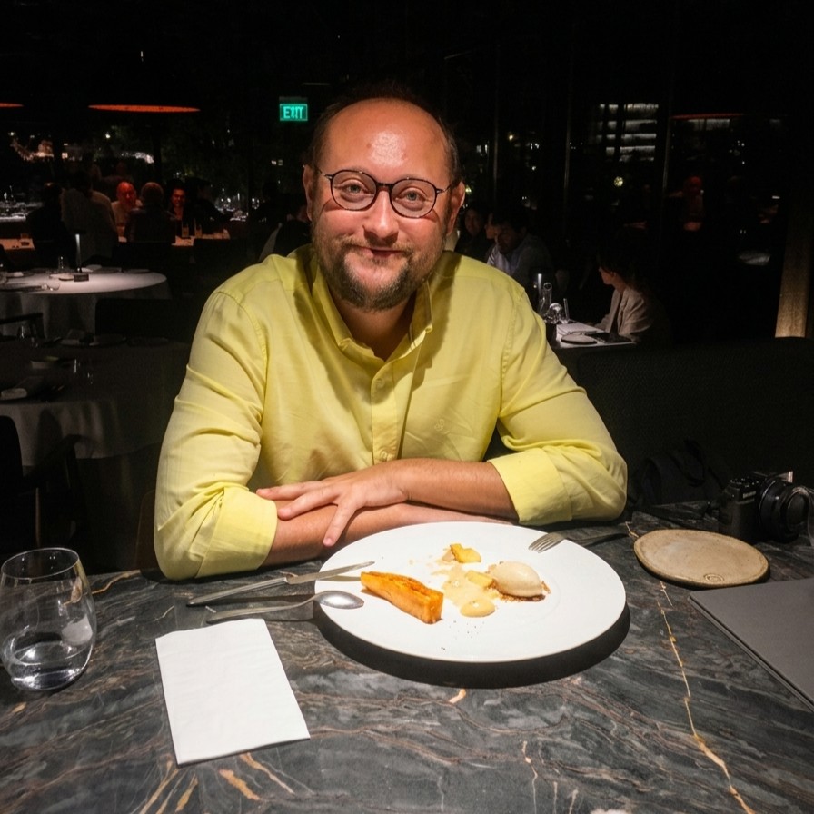 A medium shot photograph of a smiling man with round glasses and a yellow shirt, seated at a dark marble table with a white dessert plate, in a dimly lit upscale restaurant with a green 'EXIT' sign in the background.