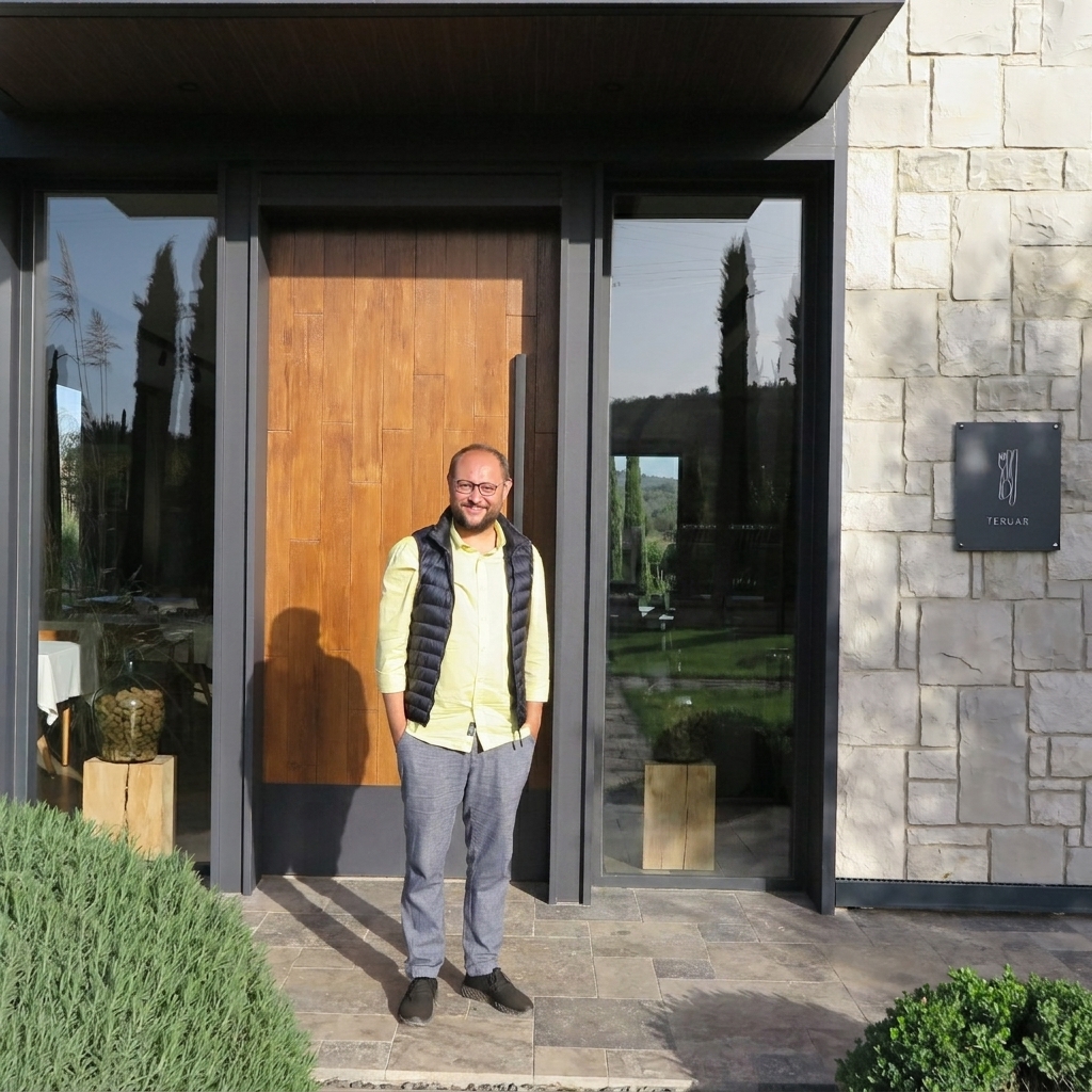 A man with glasses and a beard, wearing a yellow shirt and black vest, stands smiling with hands in his pockets outside the modern entrance of Teruar Urla restaurant. The entrance features stone walls, glass panels, a wood door, and a black plaque with the logo and text "TERUAR." Foreground greenery includes pampas grass and other bushes.
