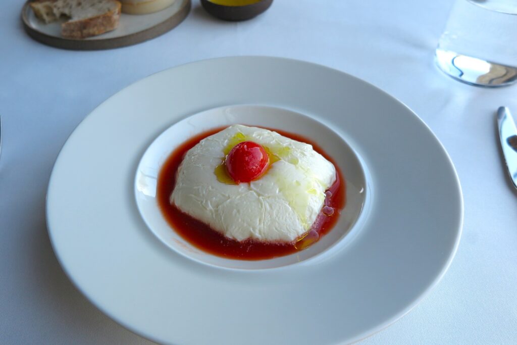 A soft white cheese mousse dish served in a pool of red tomato sauce with olive oil drizzle and a cherry tomato garnish on a white rimmed plate at Teruar Urla.