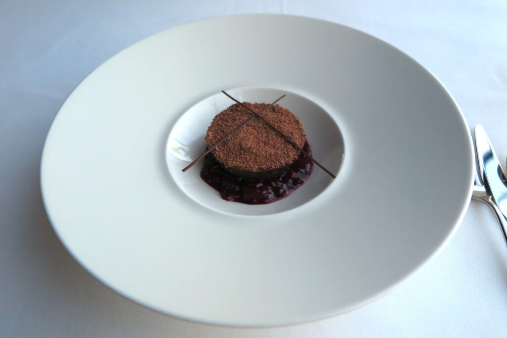A round, cocoa-dusted chocolate dessert resting on a bed of dark sour cherry compote, garnished with thin chocolate sticks, served in a wide white dish at Teruar Urla.