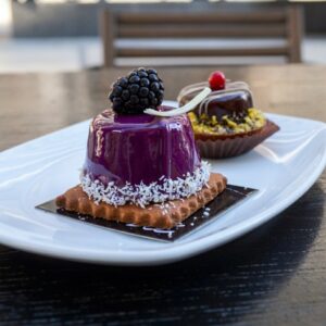 A close-up photo of two mirror-glaze mousse desserts from Blé patisserie in Thessaloniki, served on a white plate. One has a deep purple glaze topped with a blackberry and white chocolate curl, while the other has a dark chocolate glaze with pistachios and a red currant.