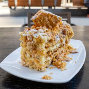 A close-up photo of a Mille-Feuille pastry served on a white ceramic plate at Blé patisserie in Thessaloniki. The dessert features crisp, golden puff pastry layers filled with smooth pastry cream and covered with puff pastry crumbs and powdered sugar, with a small pastry piece on top.
