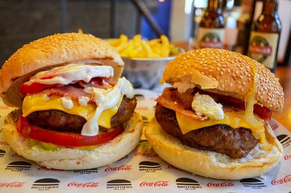 Two large, gourmet beef burgers with cheese, bacon, and toppings on sesame buns, served on branded parchment paper at Brothers In Law restaurant in Thessaloniki, with fries and beer bottles blurred in the background.