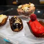 Three mini eclairs – white chocolate, dark chocolate with almonds, and raspberry glaze – sit on a glass plate, with a layered dessert bowl blurred in the background.