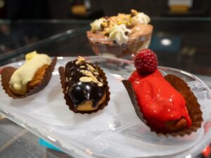 Three mini eclairs – white chocolate, dark chocolate with almonds, and raspberry glaze – sit on a glass plate, with a layered dessert bowl blurred in the background.