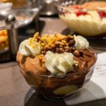 A glass bowl filled with profiteroles, chocolate sauce, and whipped cream topped with nuts, sitting on a counter at Choureal in Thessaloniki.