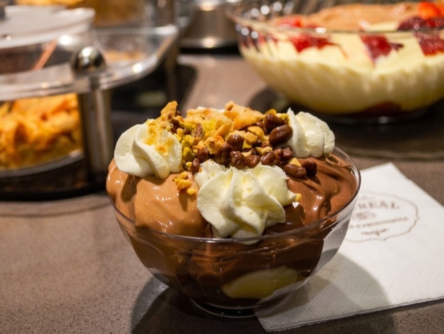 A glass bowl filled with profiteroles, chocolate sauce, and whipped cream topped with nuts, sitting on a counter at Choureal in Thessaloniki.