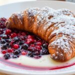 A close-up, high-angle photo of Estrella’s Bougatsan in Thessaloniki, featuring a golden-brown croissant dusted with powdered sugar, served with vanilla custard and mixed berry compote on a white plate.