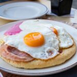 A close-up photo of Estrella’s Breakfast Pizza in Thessaloniki, served on a koulouri topped with a golden-brown slice of ham and two sunny-side-up eggs with bright, glossy yolks. A bowl of pink dip is visible on the side, and the plate sits on a wooden table.