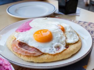 A close-up photo of Estrella’s Breakfast Pizza in Thessaloniki, served on a koulouri topped with a golden-brown slice of ham and two sunny-side-up eggs with bright, glossy yolks. A bowl of pink dip is visible on the side, and the plate sits on a wooden table.