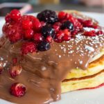 A close-up photo of three thick, fluffy pancakes topped with chocolate sauce, mixed berries, and powdered sugar, served on a white plate. The background is softly blurred.
