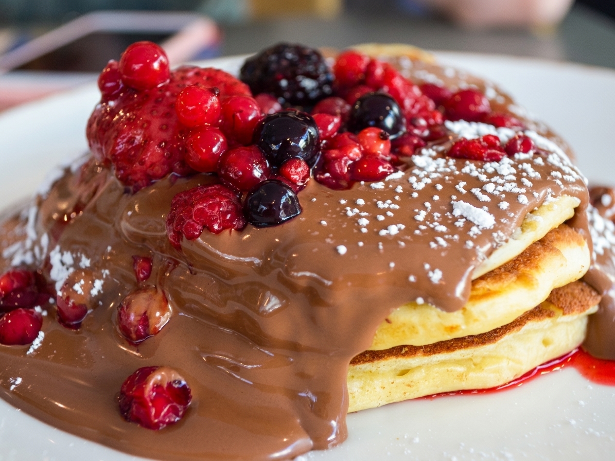 A close-up photo of three thick, fluffy pancakes topped with chocolate sauce, mixed berries, and powdered sugar, served on a white plate. The background is softly blurred.