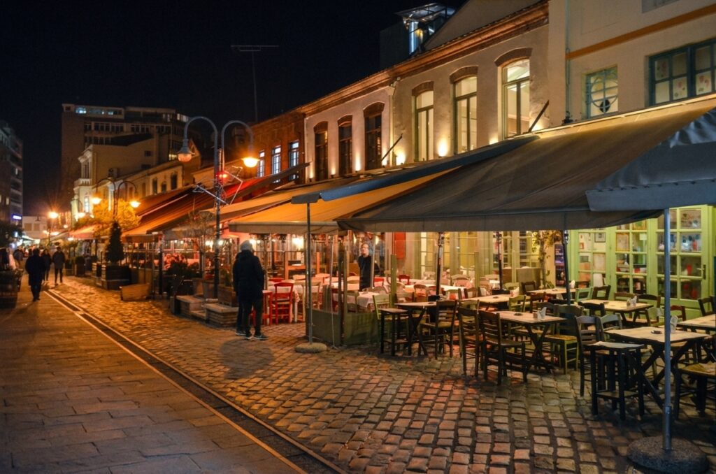 A night view of a cobblestone pedestrian street in Thessaloniki's historic Ladadika district, with warmly lit tavernas and outdoor seating.