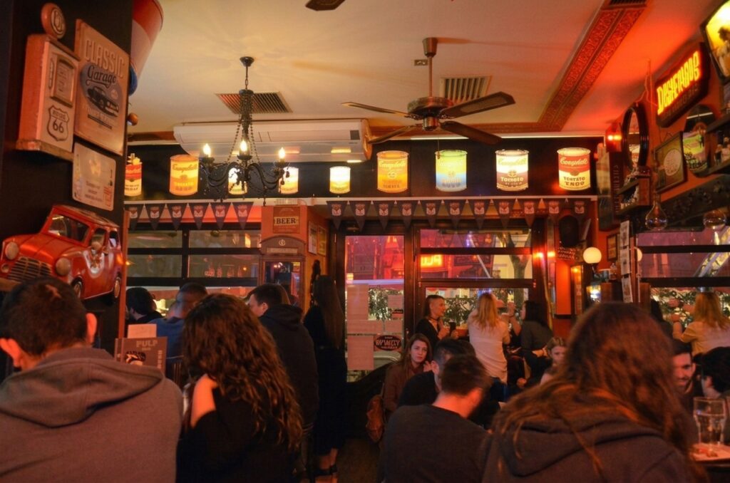 A vibrant photograph of the interior of The Pulp Bar in Thessaloniki, packed with patrons under warm red and amber lighting. The decor is eclectic, featuring retro signs like 'Classic Garage,' a vintage red car front, and a unique row of lit Campbell's soup can lamps.
