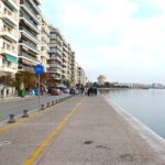 A wide-angle view of the Thessaloniki waterfront, with the Thermaikos Gulf on the right, city buildings on the left, and the White Tower in the background. A yellow-lined bicycle path and blue bicycle sign are visible in the foreground.