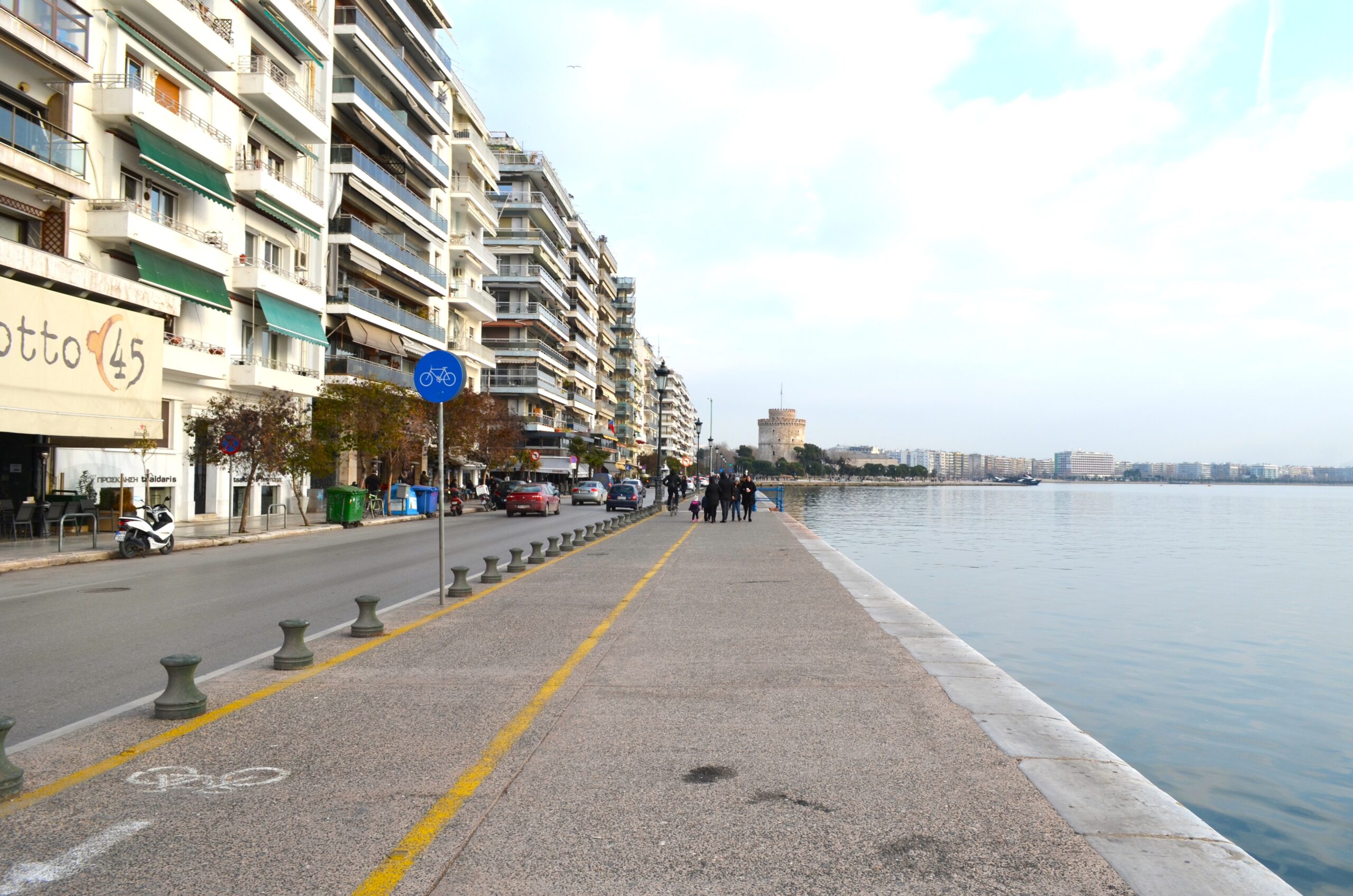 A wide-angle view of the Thessaloniki waterfront, with the Thermaikos Gulf on the right, city buildings on the left, and the White Tower in the background. A yellow-lined bicycle path and blue bicycle sign are visible in the foreground.